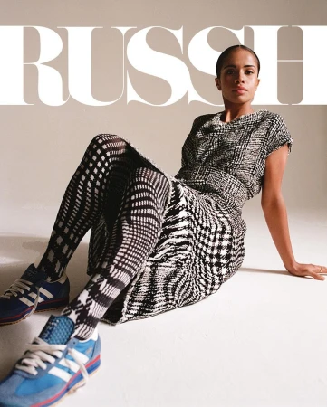 Model seated on white studio floor wearing black and white plaid dress, patterned tights, and blue sneakers.