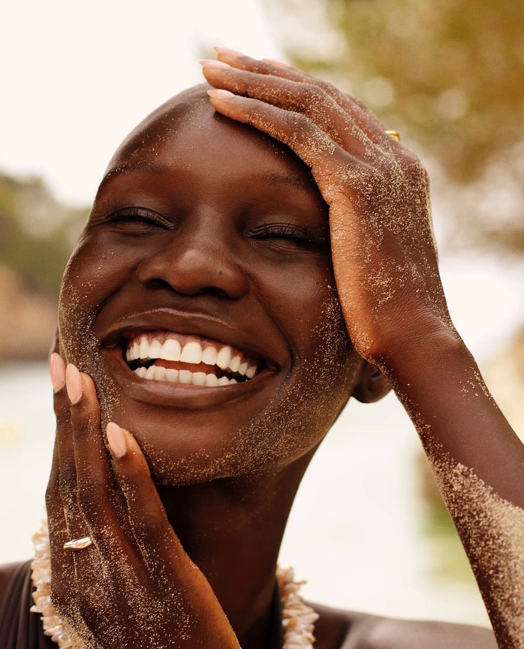 Close-up portrait of a smiling, dark-skinned woman with sand on her face and hands, wearing a shell necklace and ring.