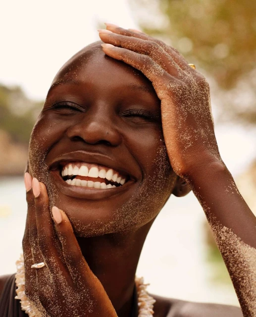 Close-up portrait of a smiling, dark-skinned woman with sand on her face and hands, wearing a shell necklace and ring.