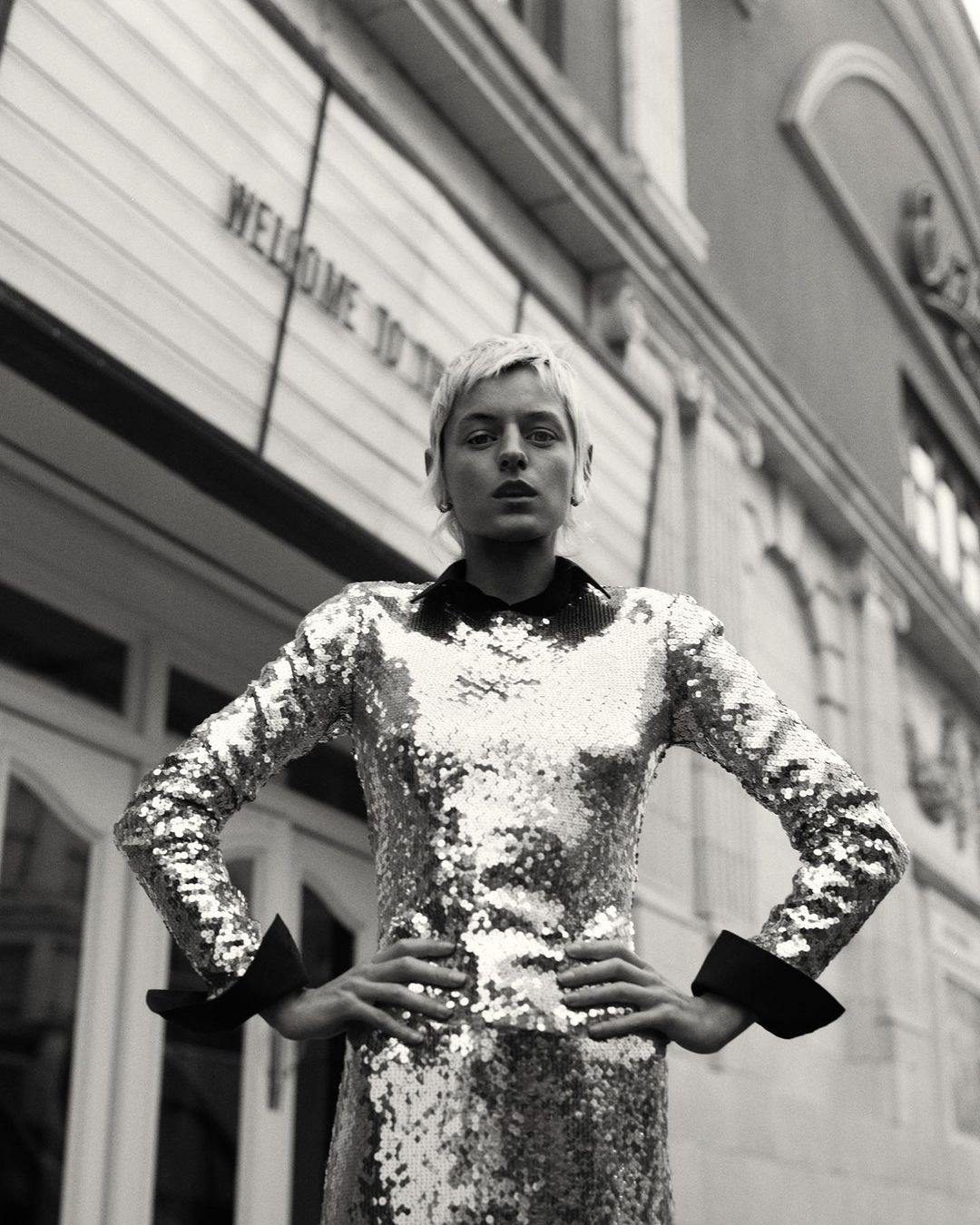 Monochrome portrait of a woman with short blonde hair wearing a sequined long-sleeve dress with hands on hips outside a building