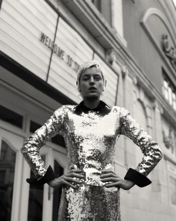 Monochrome portrait of a woman with short blonde hair wearing a sequined long-sleeve dress with hands on hips outside a building
