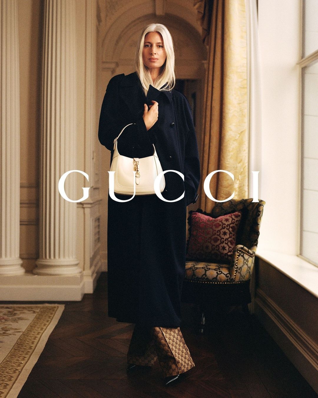 Woman with long blonde hair wearing a dark coat and holding a white handbag inside a grand room