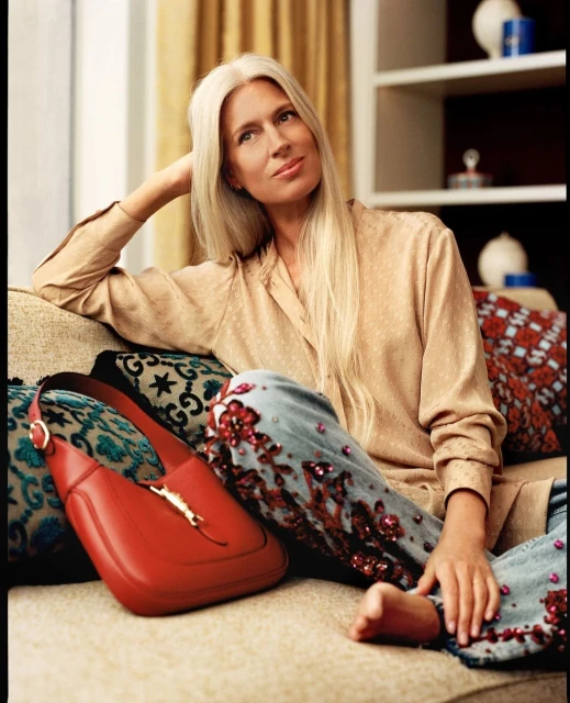 Elegant woman sits on couch with purse, wearing embellished jeans and a blouse. Natural light floods the serene interior.