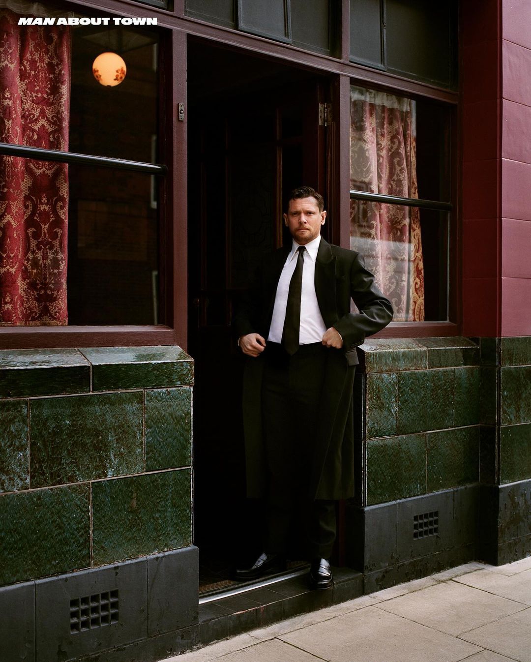 Portrait of a man in a dark suit posing in a doorway with an interior view featuring patterned curtains and a hanging light fixture.