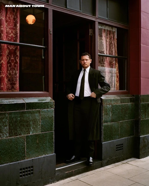 Portrait of a man in a dark suit posing in a doorway with an interior view featuring patterned curtains and a hanging light fixture.