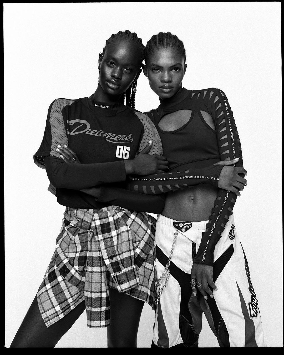 Black and white studio shot of two athletic female fashion models in sporty separates, hugging, against a bright white backdrop.
