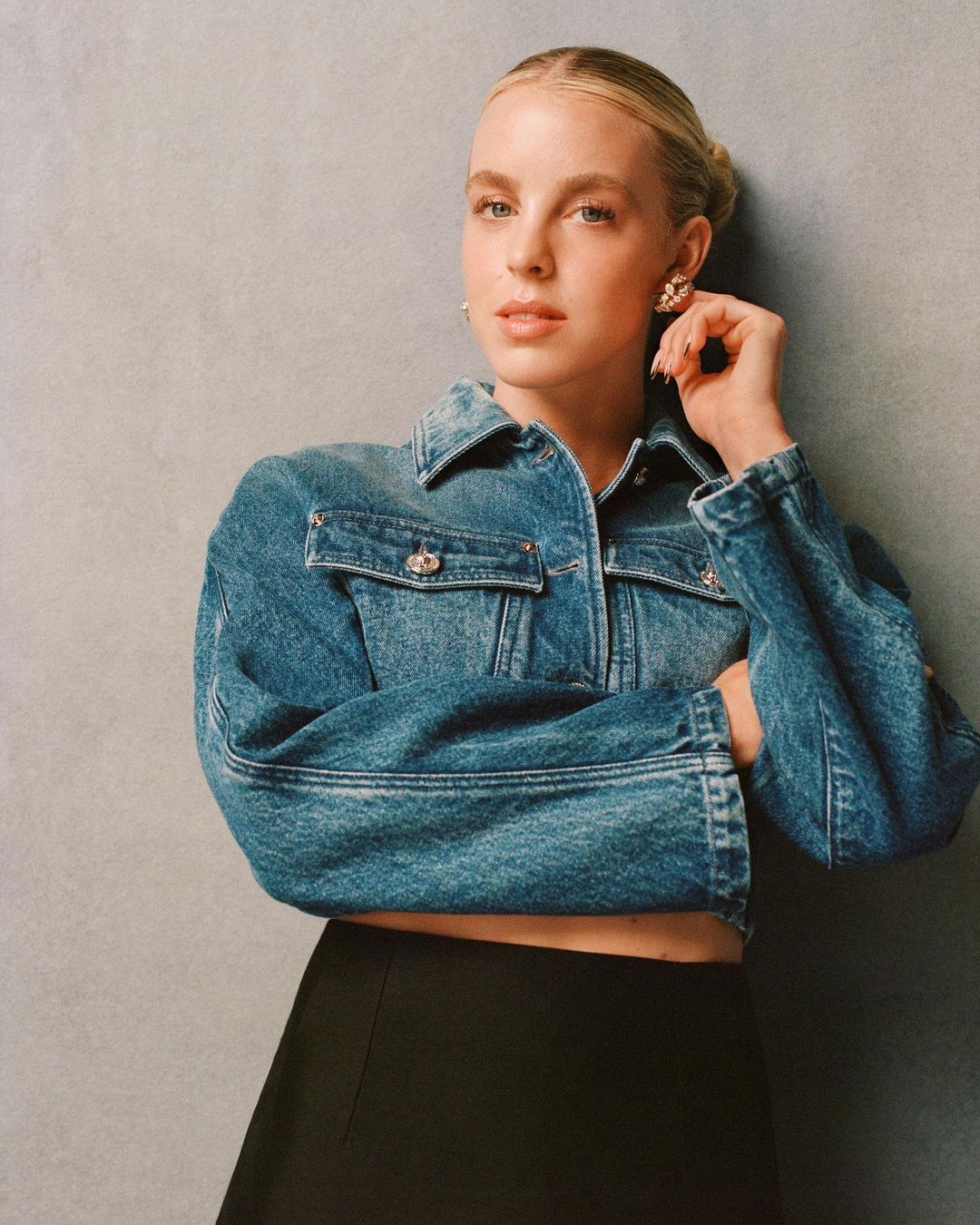 Blonde woman poses in a denim jacket and black skirt against a grey backdrop, showcasing a modern and stylish look in a studio setting.