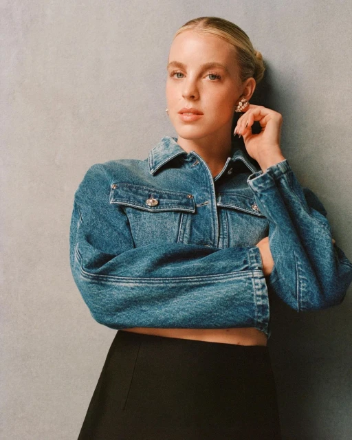 Blonde woman poses in a denim jacket and black skirt against a grey backdrop, showcasing a modern and stylish look in a studio setting.