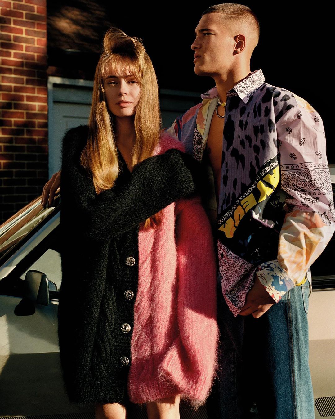 A woman in a split black and pink fuzzy cardigan and a man in a patterned shirt standing next to a car