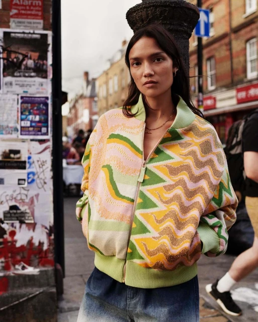 A fashionable woman in a patterned zip-up jacket stands on a city street near buildings and posters, exuding confidence in daylight.