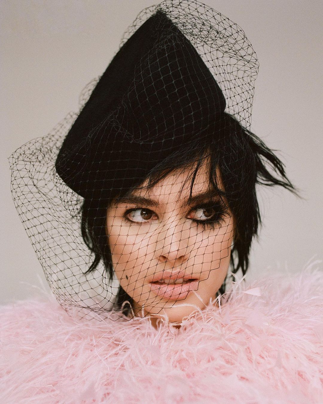 Close-up of a woman wearing a black hat with a net veil and a pink feathered boa. She has dark hair and is looking to the side.