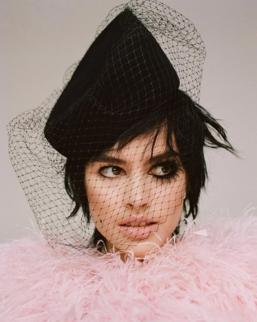 Close-up of a woman wearing a black hat with a net veil and a pink feathered boa. She has dark hair and is looking to the side.