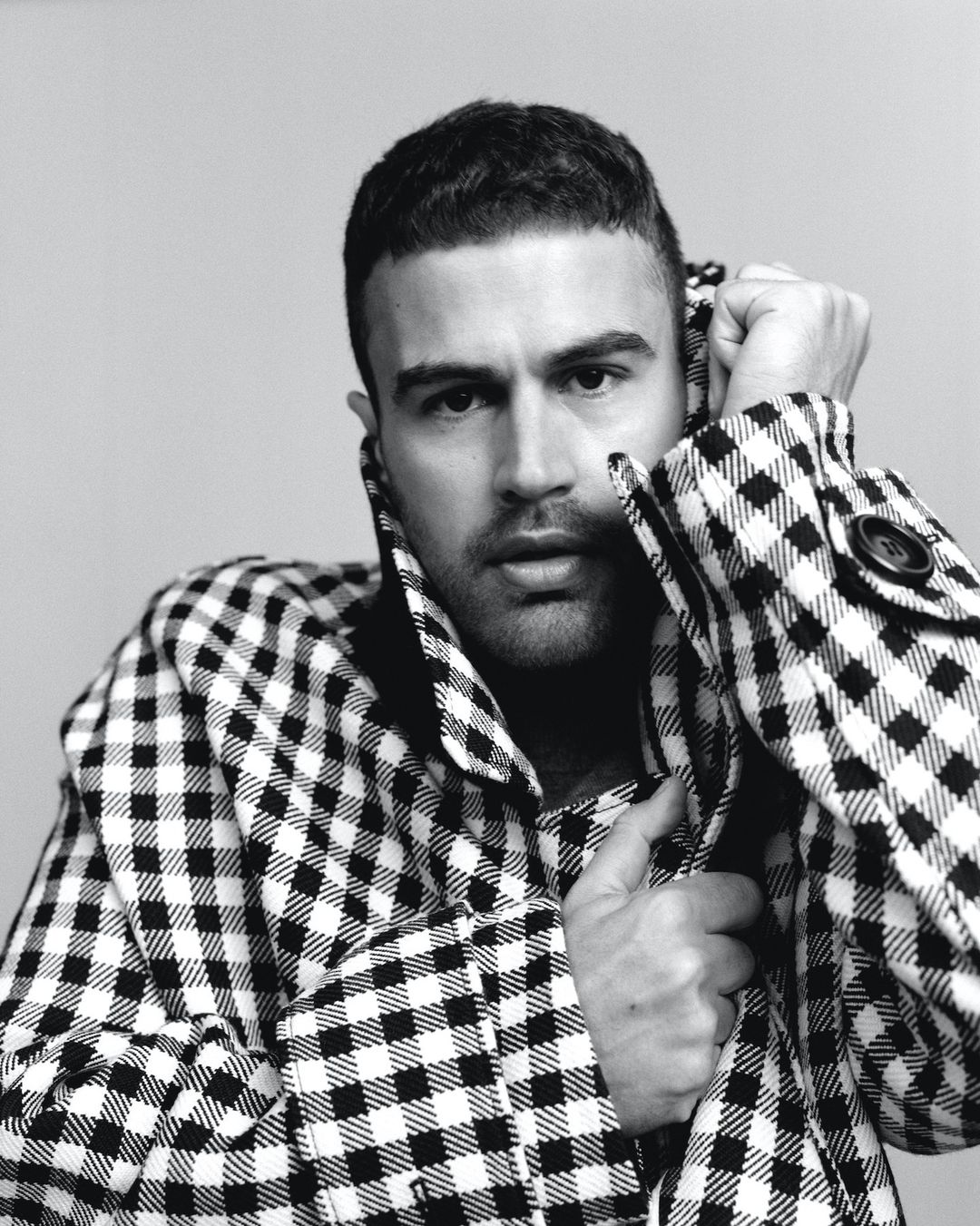 Portrait of a man in a black and white gingham patterned coat holding the collar up against a light grey background