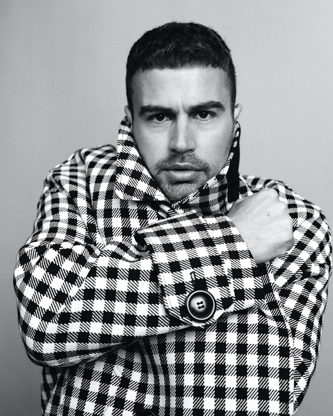 Monochrome portrait of a man in a patterned coat, gazing into the camera with a neutral expression against a light gray backdrop.