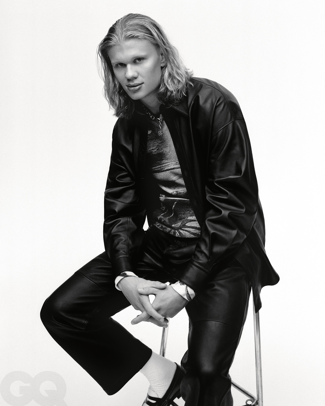 Black and white portrait of a blonde male model in a shiny shirt and black leather jacket, seated on a silver stool in a photo studio.