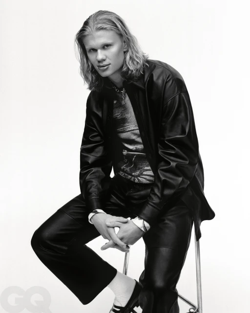 Black and white portrait of a blonde male model in a shiny shirt and black leather jacket, seated on a silver stool in a photo studio.