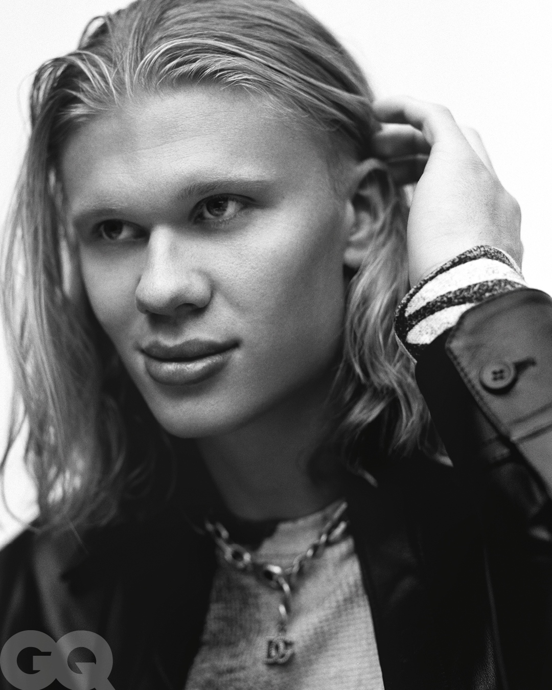 Close-up portrait of a man with long blond hair touching his hair, wearing a dark jacket and chain necklace, photographed by Buzz White
