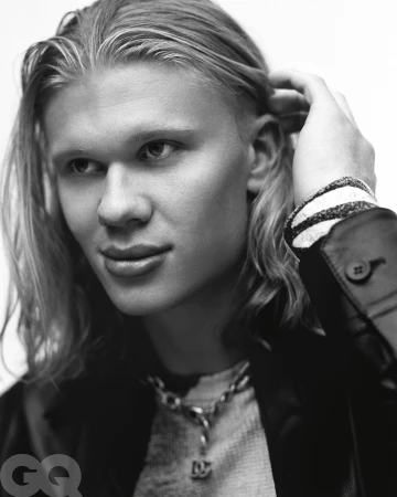 Close-up portrait of a man with long blond hair touching his hair, wearing a dark jacket and chain necklace, photographed by Buzz White