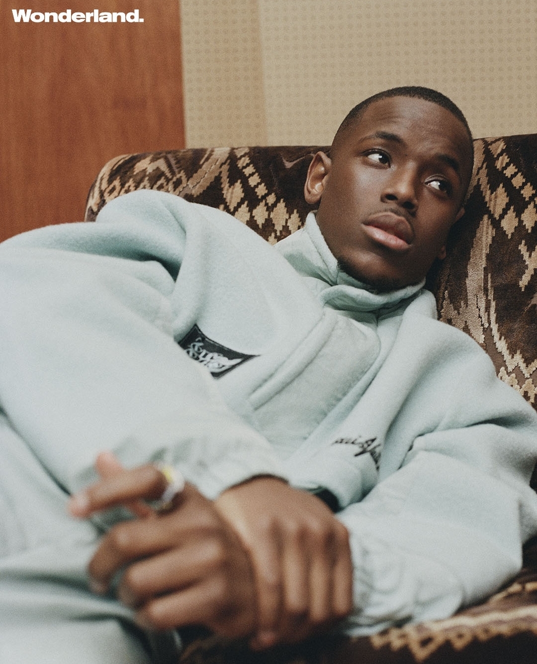 Portrait of a Black male model in light blue sweatsuit relaxing on patterned chair in an indoor setting with soft, diffused lighting.