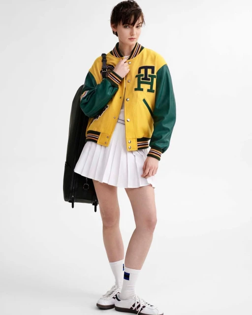 Full-length shot of an adult female in a varsity jacket and white pleated tennis skirt, holding a black case, against a white background.