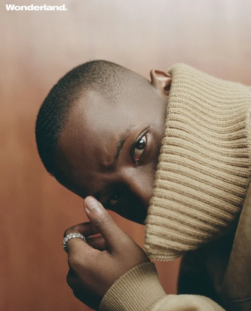 Close-up portrait of a Black male wearing a beige turtleneck sweater and a diamond ring against a warm orange-brown background.