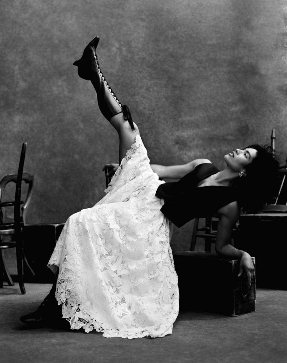 A woman in a black top and white lace skirt poses with her leg raised in black lace-up boots against a mottled background.