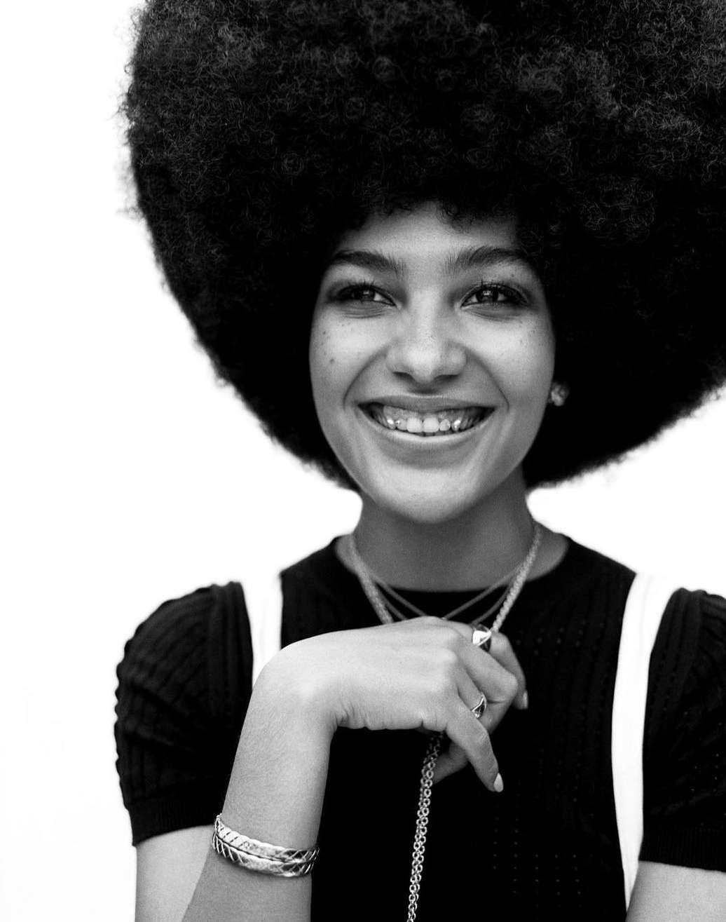 Close-up black and white portrait of a young woman smiling, featuring her natural hair, silver jewelry, and braces.