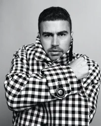 Man wearing a black and white checkered coat posing against a plain grey studio backdrop