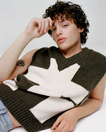 Young man with dark curly hair wearing an olive green and white sweater vest against a white background