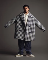 Man wearing oversized houndstooth coat and striped trousers standing against grey background