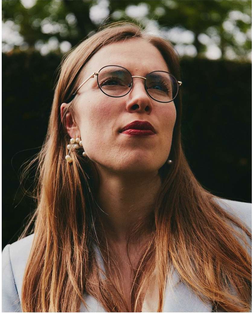 Portrait of a woman with long auburn hair wearing glasses, a light blue blazer, and pearl earrings, gazing upward against a dark green background.