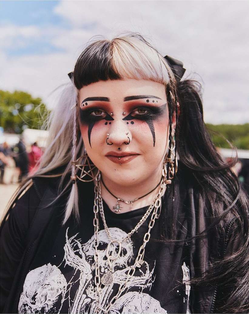 A young adult woman with striking dark makeup and split-dye hair poses in an outdoor setting, showcasing an alternative fashion style.