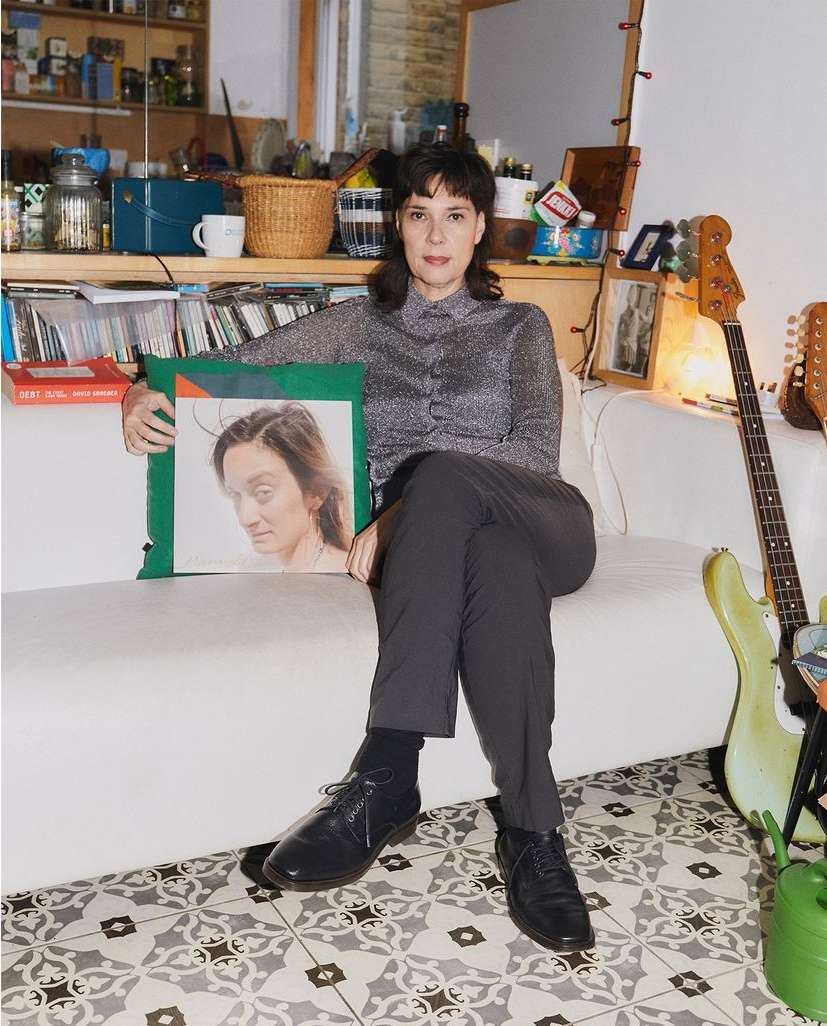 Indoor portrait of an adult woman sitting on a white couch holding a large portrait of a face, with guitars in the background.