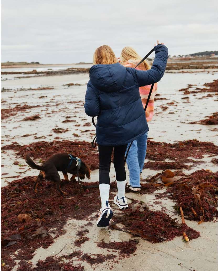 Two children walk with a brown dog on a sandy beach with patches of seaweed under a cloudy sky, holding a black dog leash.