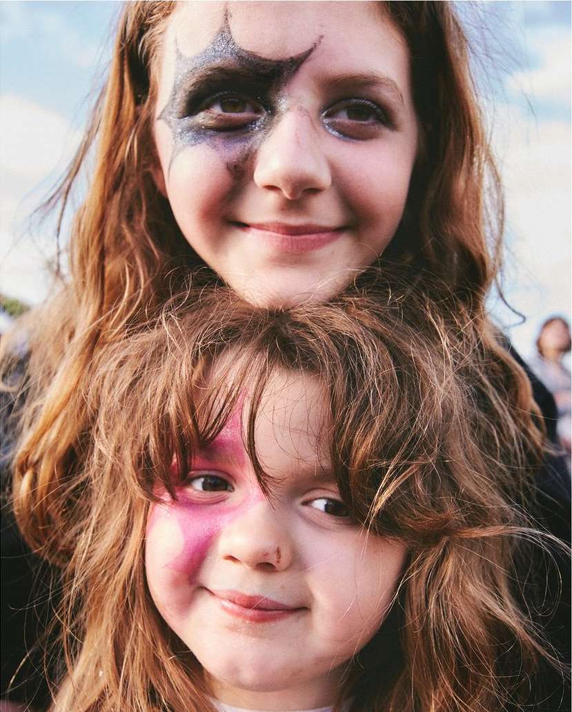 Two smiling girls with painted faces pose outdoors against a bright sky. The children have long red hair and happy expressions.