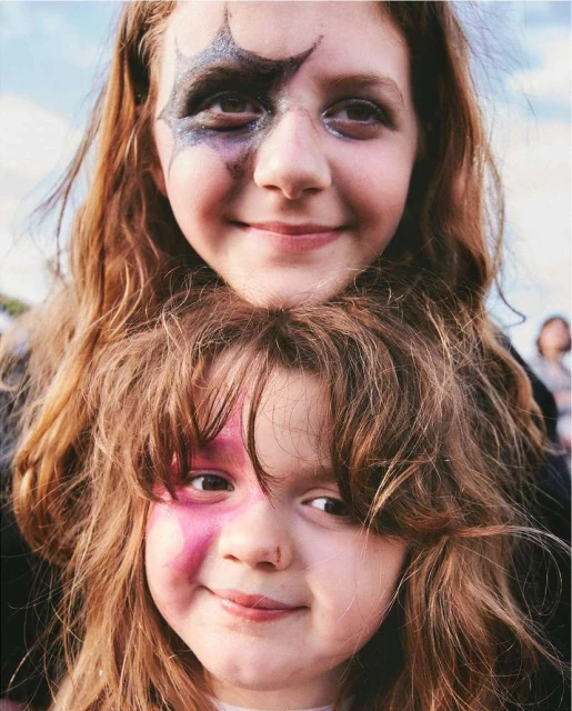 Two smiling girls with painted faces pose outdoors against a bright sky. The children have long red hair and happy expressions.
