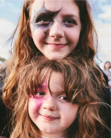 Two smiling girls with painted faces pose outdoors against a bright sky. The children have long red hair and happy expressions.