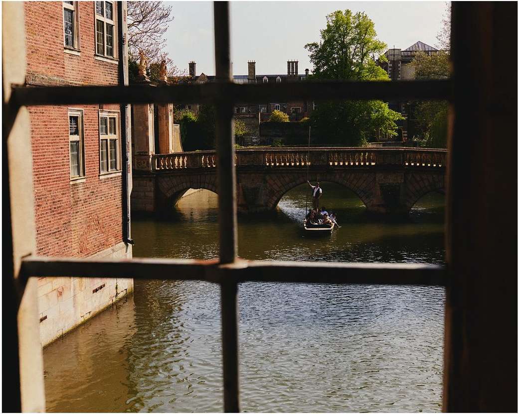 Scenic view through window of punting on river Cam, Cambridge, England; includes boat, bridge, architectural buildings and natural landscape.