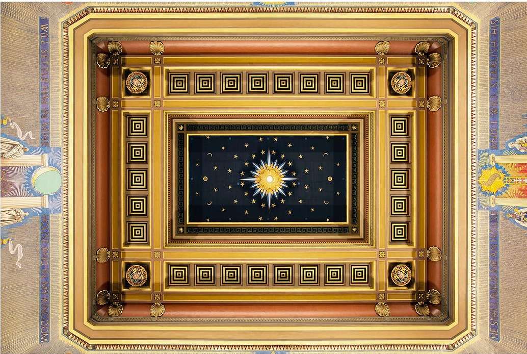 Ornate ceiling with a central sunburst design surrounded by geometric patterns, detailed molding, and decorative elements in gold and brown tones.