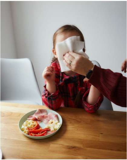 Young child with red plaid shirt at a wooden table, having their face cleaned with a white tissue by an adult at an indoor dining setting.