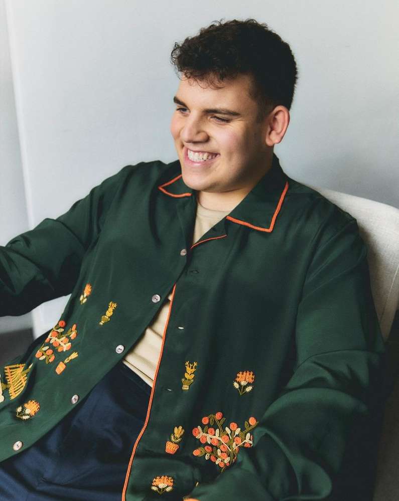 Man smiles in a portrait, wearing a dark green shirt with orange trim and floral embroidery. The photo is set against a light gray wall.
