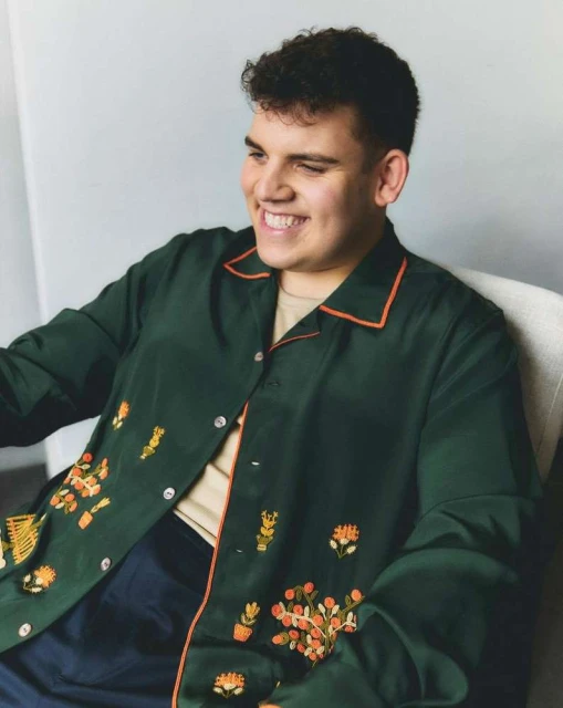 Man smiles in a portrait, wearing a dark green shirt with orange trim and floral embroidery. The photo is set against a light gray wall.
