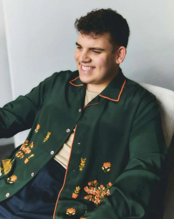 Man smiles in a portrait, wearing a dark green shirt with orange trim and floral embroidery. The photo is set against a light gray wall.