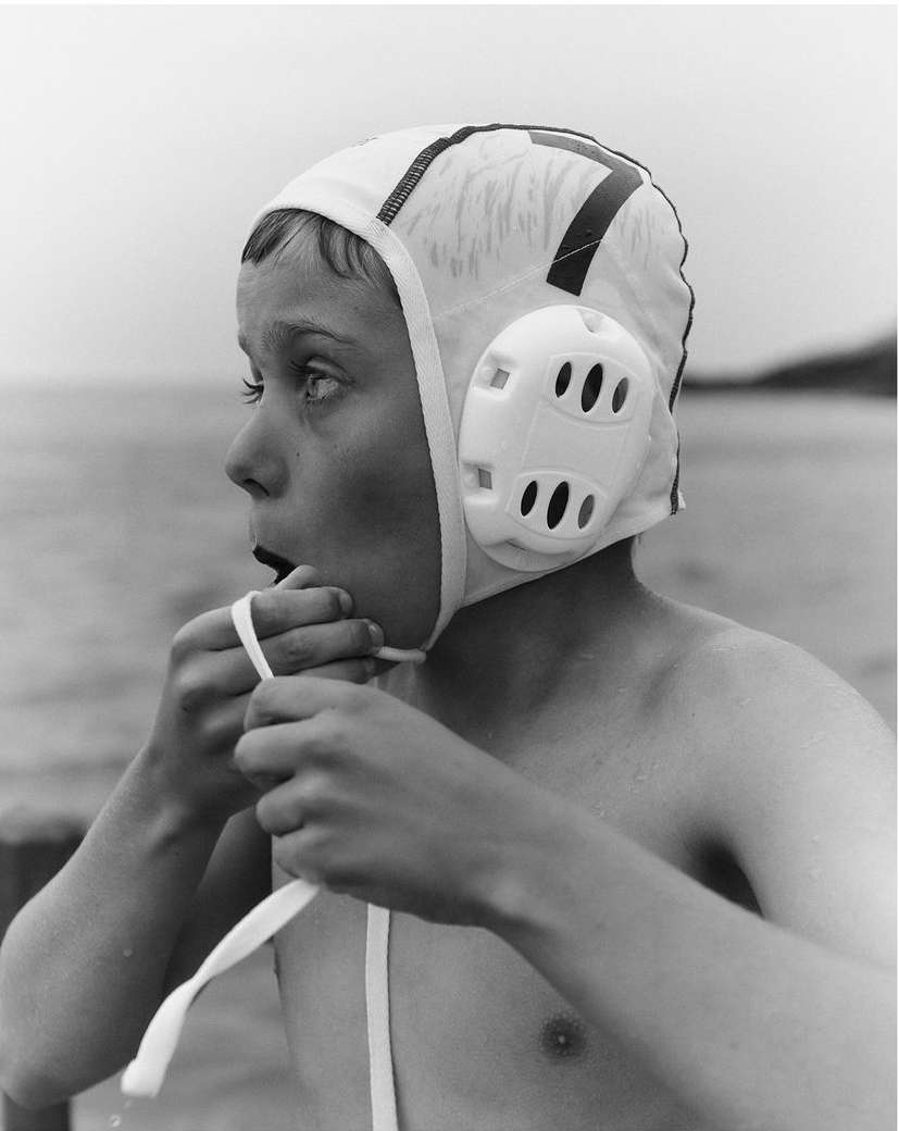 Boy with water polo cap ties the strings, looking to the left, with water and trees visible in the background, in black and white.