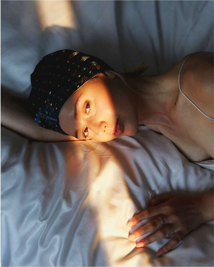 Woman with headscarf lying on white silk bedsheets, lit by warm sunlight, creating shadows and highlights on face and bedding.