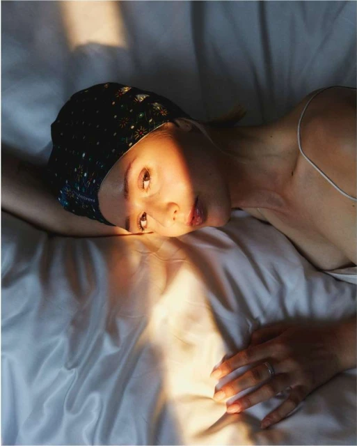 Woman with headscarf lying on white silk bedsheets, lit by warm sunlight, creating shadows and highlights on face and bedding.