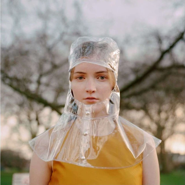 Woman in a yellow dress and a transparent plastic rain bonnet stands outdoors with a blurred tree background, portrait view.