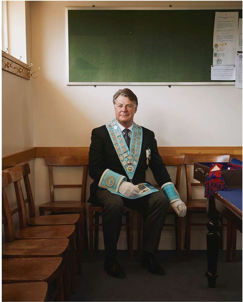 Adult male seated indoors, wearing a suit and ceremonial garb, including a sash, medallions, and gloves, in a well-lit room.