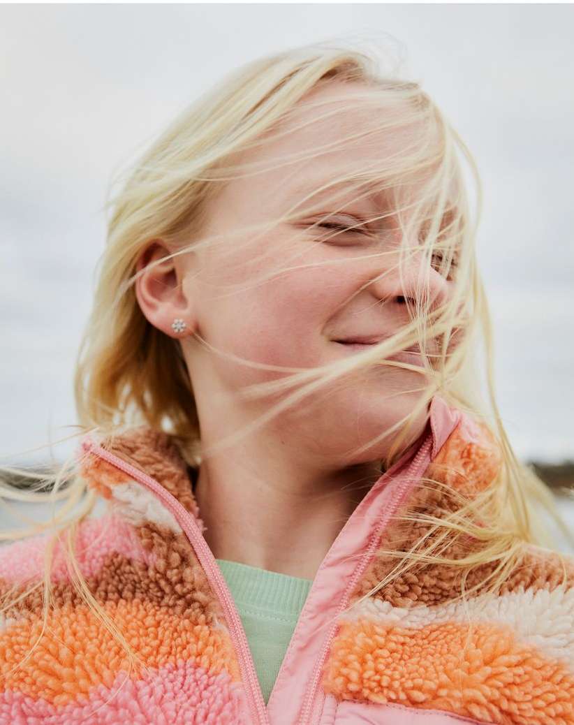 Close-up of a young girl with blonde hair and closed eyes, wearing a pink and orange jacket, against an overcast sky background.