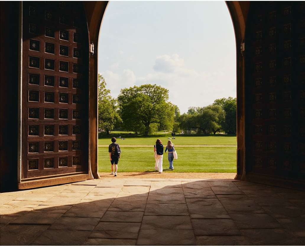Three figures walk through an arched doorway leading to a green field with trees under a bright sky, framed by a stone floor.